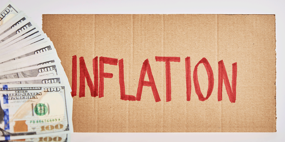 Inflation written in red on a piece of cardboard next to a stack of $100 bills.
