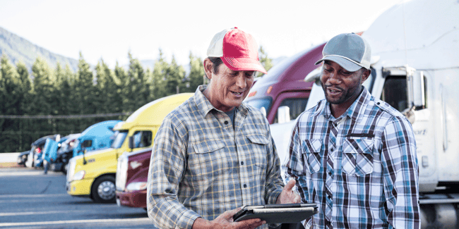 Two truck drivers working together while they review something on a tablet screen. They stand in front of a row of parked semi-trucks; ATS
