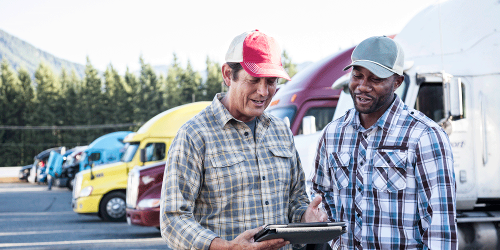 Two truck drivers working together while they review something on a tablet screen. They stand in front of a row of parked semi-trucks.