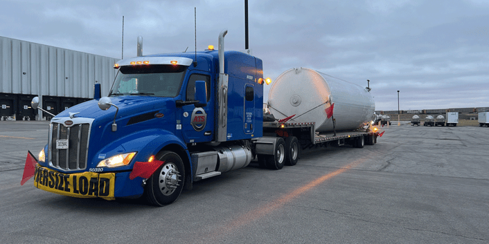 Blue ATS flatbed truck with yellow oversize load banner. A large tank is secured to the trailer, Anderson Trucking Service, company truck driver positions
