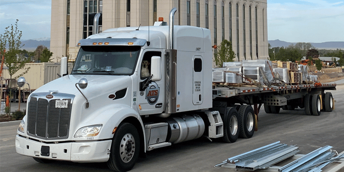 A white semi-struck with an attached flatbed trailer. A dog sits in the driver's seat, ATS company truck driver positions