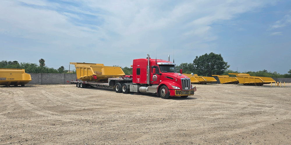 ATS truck with oversize load banner in a gravel yard surrounded by large yellow construction equipment.