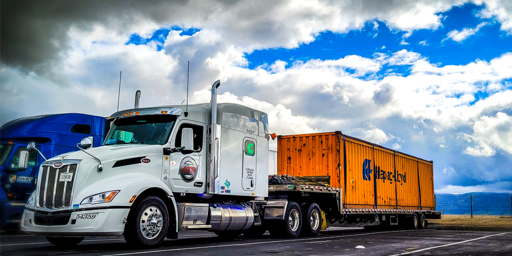 White tractor hauling an orange shipping container. The sky is bright blue and cloudy.