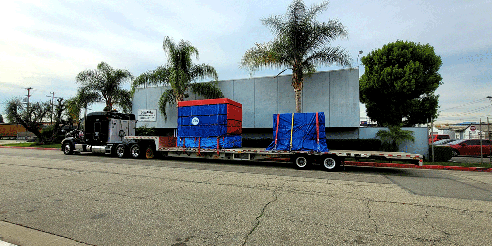 Two tarped objects on a step-deck trailer. The truck is parked in front of palm trees.