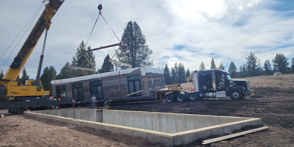 A black ATS truck with a small building structure on its trailer. A crane is about to lift the freight. An empty foundation on a dirt-covered site is next to it.