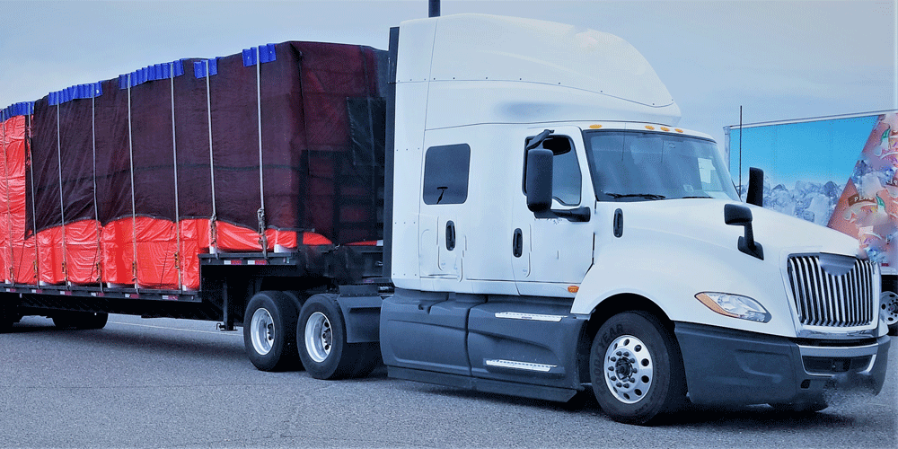 White semi-truck hauling a step-deck trailer covered in a black and red tarp.