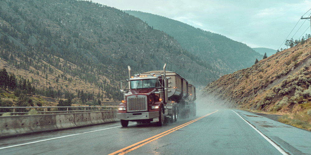 Semi-truck hauling a belly dump trailer through a hilly region. The road is wet and the tires are spraying up water from the road.