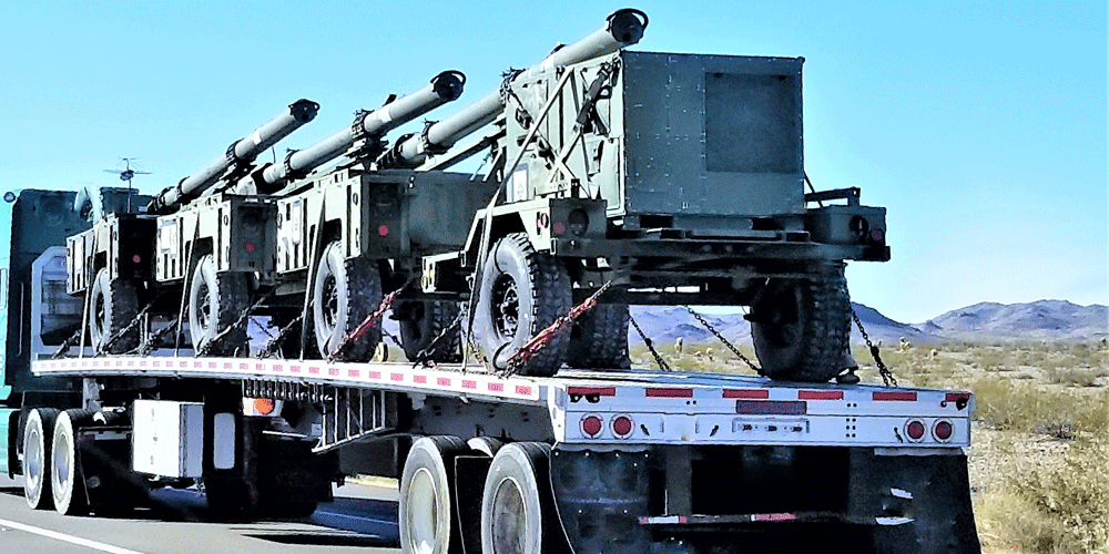 Semi-truck hauling military cannons on a flatbed trailer.
