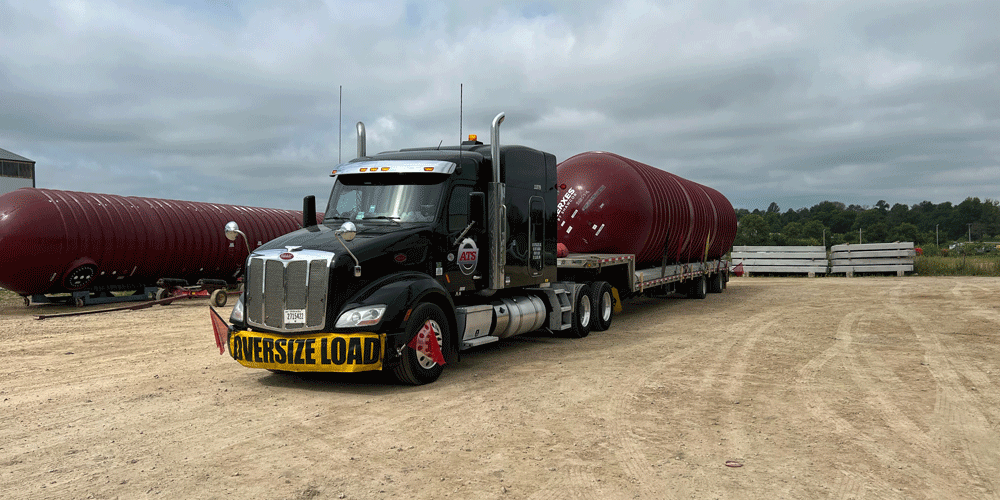 Black ATS semi-truck with oversize load banner hauling a step deck trailer with a red tank on it.