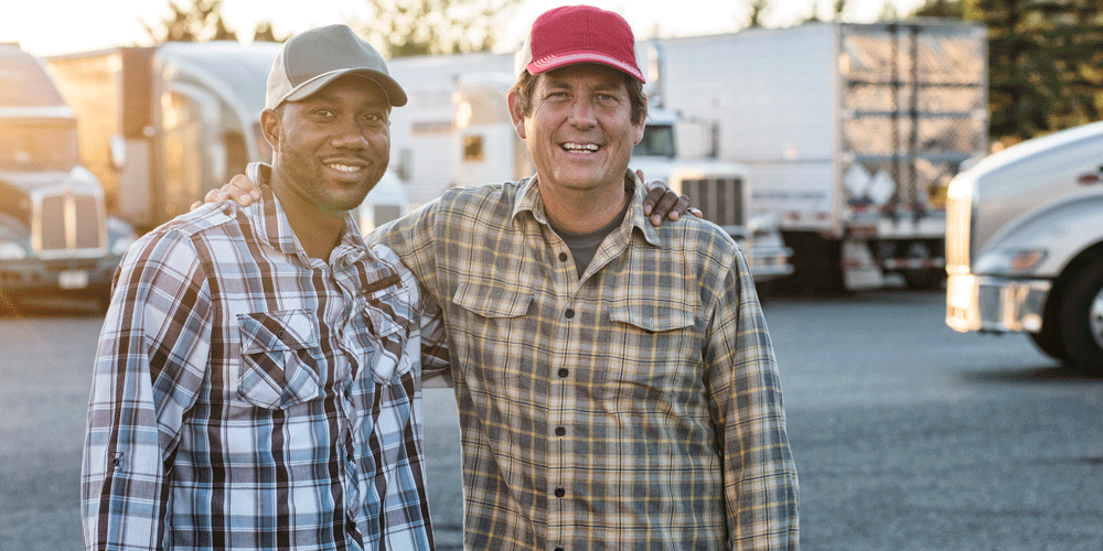 Two male truck drivers. They both wear trucker hats and button down flannel shirts.