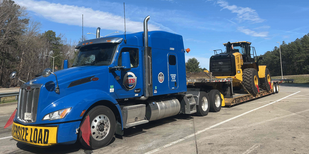 Blue ATS tractor with oversize load banner and flags hauling construction equipment.