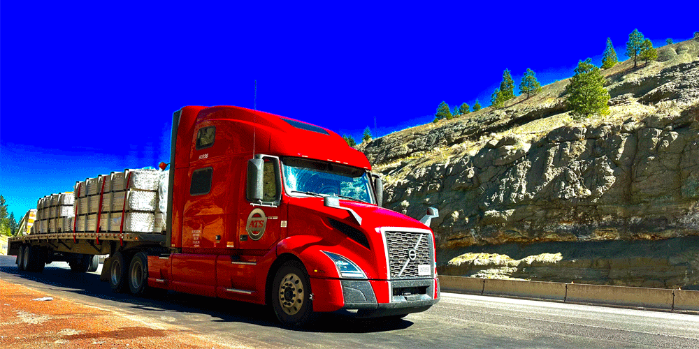 Red semi-truck hauling a flatbed in front of a rock formation.