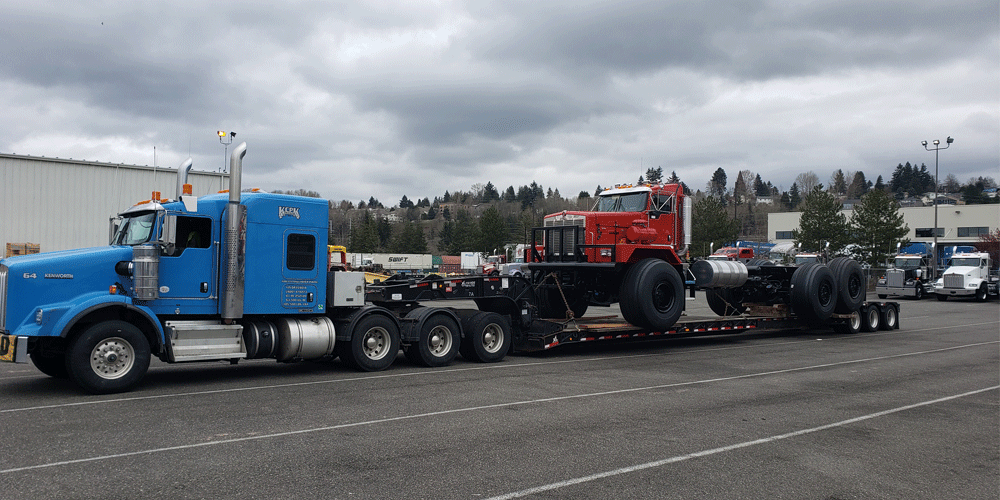 Blue semi-truck hauling smaller red truck on flatbed trailer.