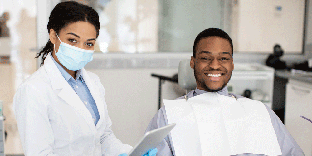 A man sitting in a dentist's chair as the dentist stands nearby in a mask.