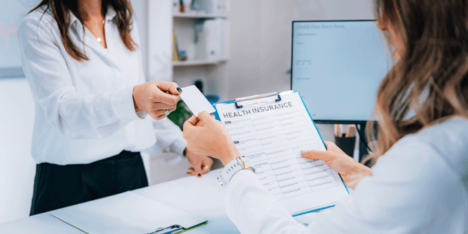 Woman handing a receptionist behind a desk and computer a card and health insurance form.