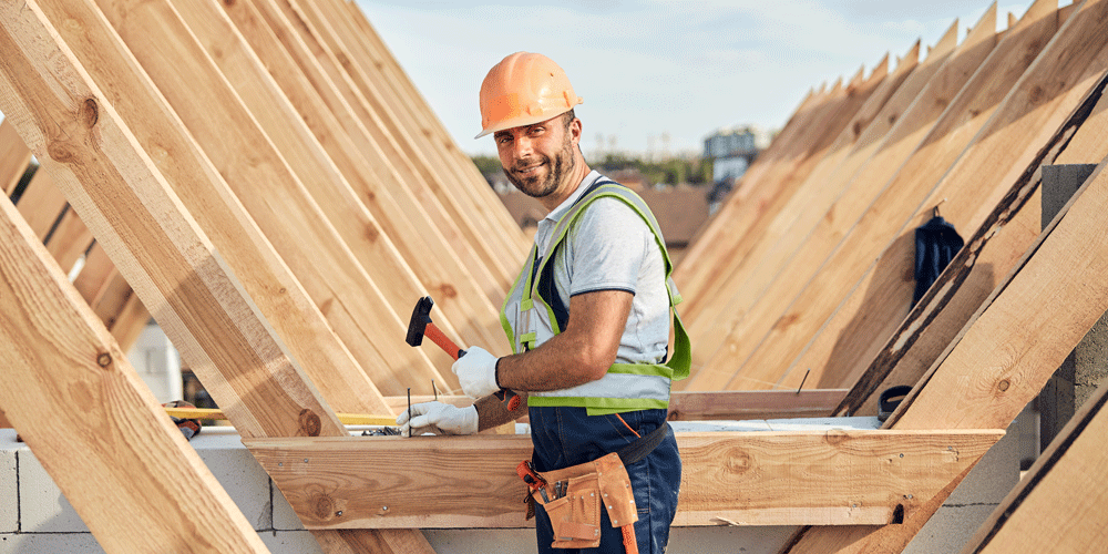 Smiling roofer in hard hat hammering nails into a roof.