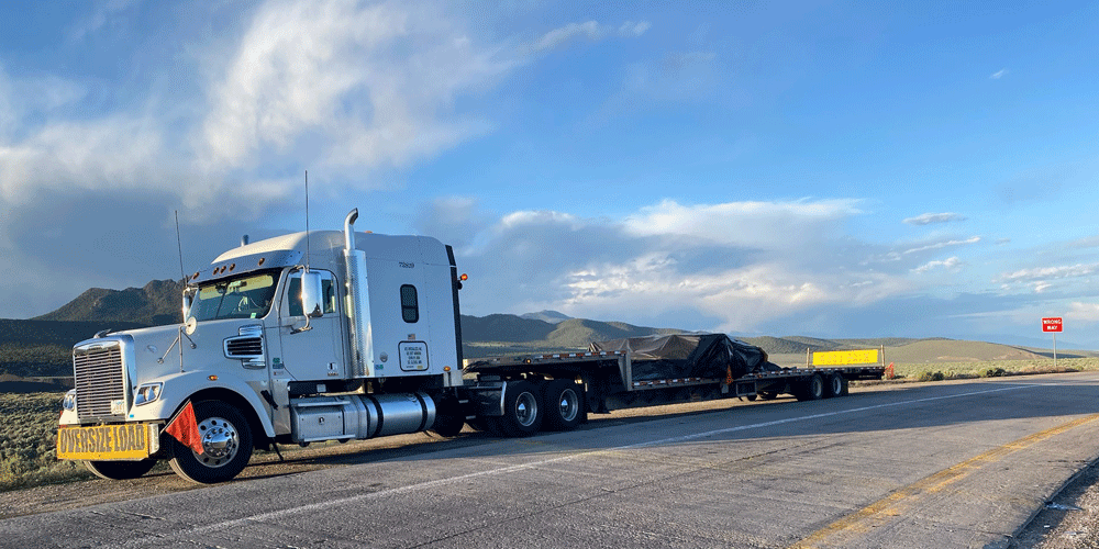 White truck with yellow oversize load banner and red flags on the front bumper. The truck hauls a tarped step deck load on a road in front of black mountains.
