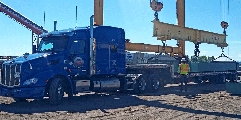 Blue truck with step-deck trailer. Freight is being loaded onto the trailer.