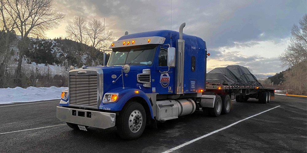 Blue semi-truck with tarped flatbed load driving on snow-covered roads.