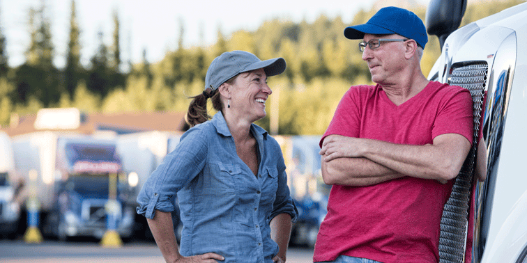 A man and woman truck driver facing each other and smiling. He leans again the grill of a semi-truck