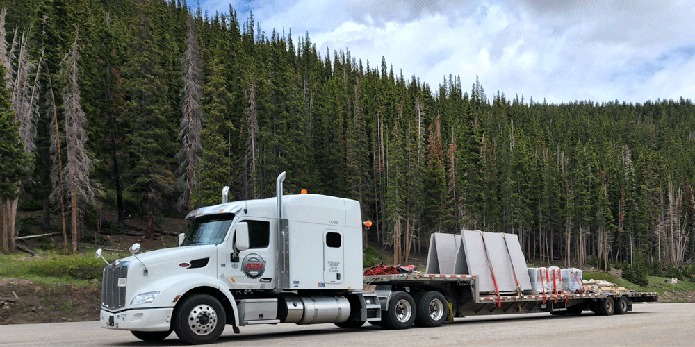 Step deck trailer loaded with granite. The semi-truck and trailer are moving in front of a forest of trees.