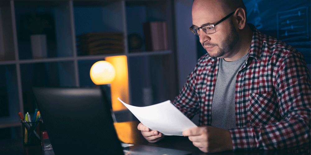 A man in a dimly lit room sitting at a desk. A laptop is in front of him and he reviews a sheet of paper in his hands.