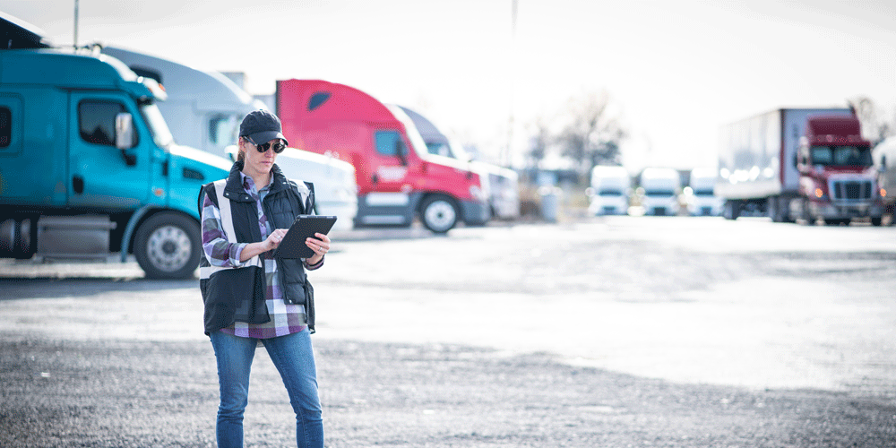 A woman in a vest, hat, and flannel shirt standing in a parking lot full of trucks. She holds a tablet.
