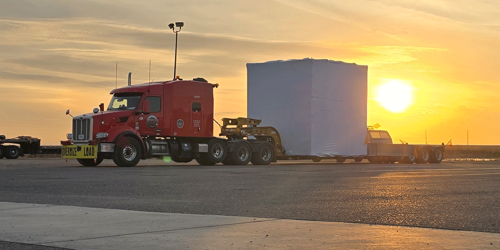 Semi-truck hauling a large electrical box as the sun sets.