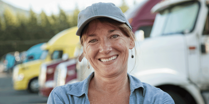 Woman truck driver smiles in front of a fleet of semi trucks; truck driver turnover, ATS