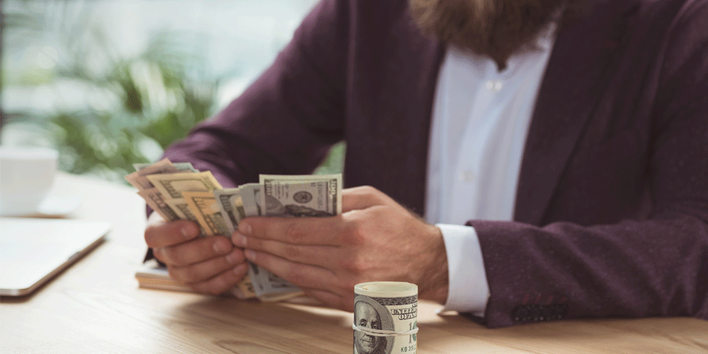 Bearded man counting $100 bills t a table.