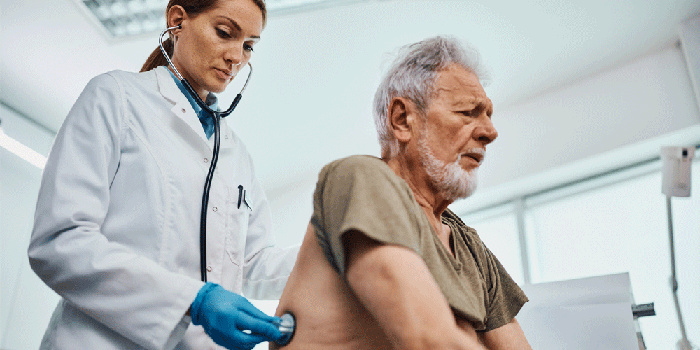 Older bearded man in a doctor's office. The doctor has her stethoscope on his back to listen to his breathing.