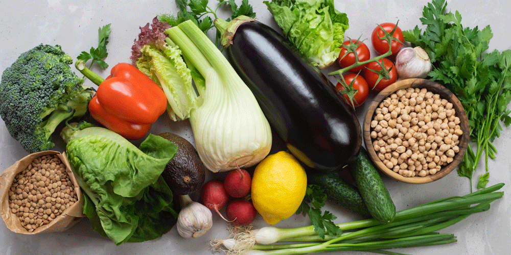 A pile of fresh produce, including on the vine tomatoes, an eggplant, greens, legumes and garlic.