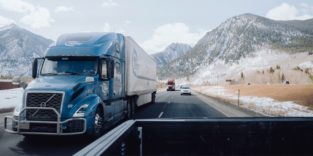 The view from the cab of a pickup truck driving through the Colorado mountains on a two-lane highway. There is a blue semi-truck and van trailer close by.