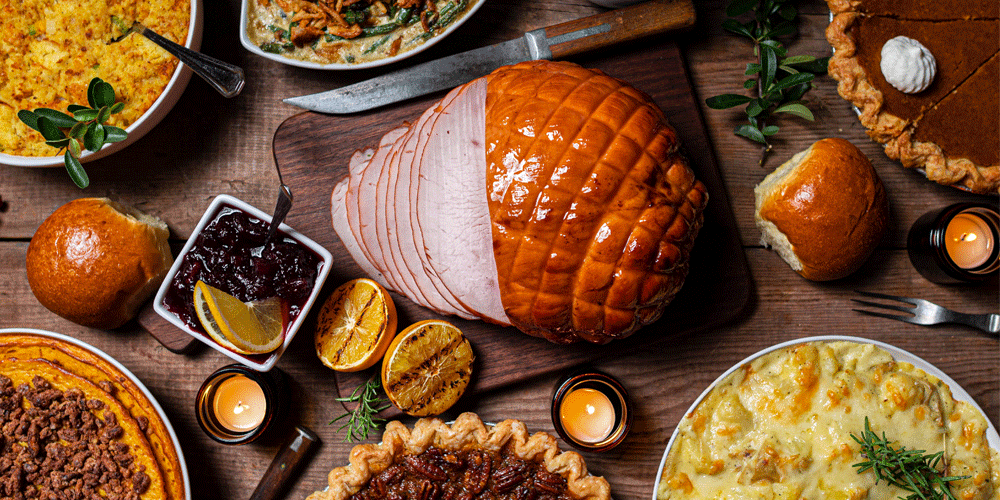 A flat lay of holiday food. There are several pies, rolls, side dishes, and a large partially sliced ham in the center.