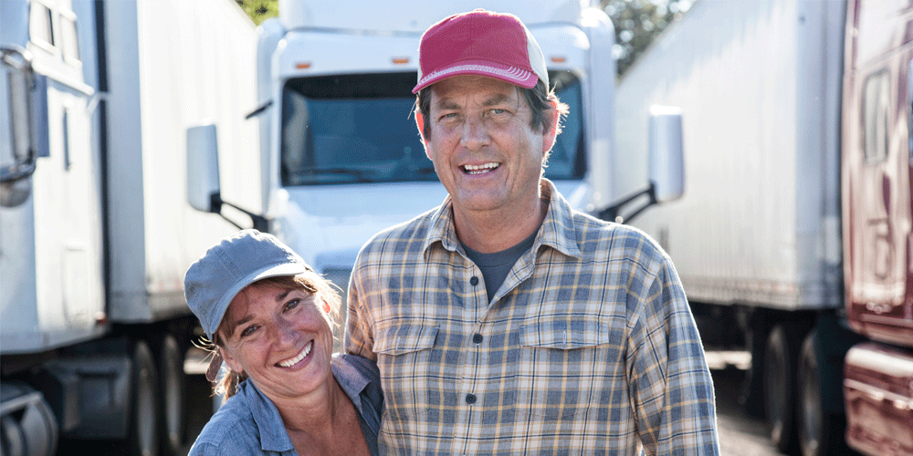 A man and wife team hug each other and smile while standing in front of several parked trucks.