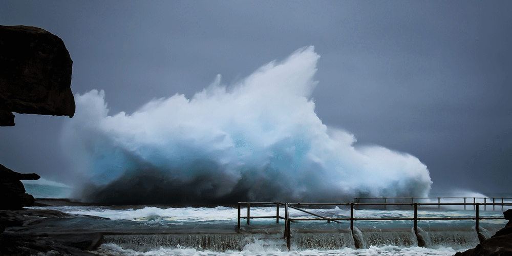 Huge ocean wave crashing over a large barrier and flooding the area.