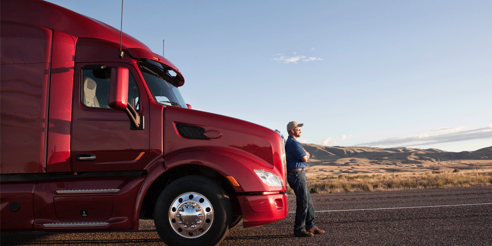 Driver leaning on red truck while parked on the side of the road in a desert landscape.