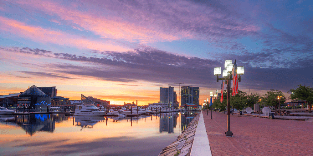 Baltimore at sunset, with a view of the harbor the focal point of the photo. The harbor is on the left with several personal yachts and on the right is a brick paved path.
