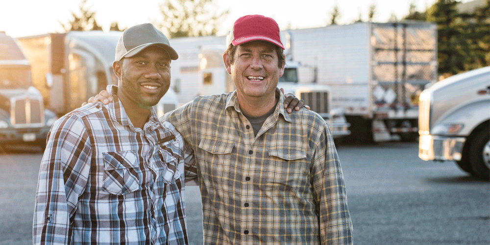 A team of two truck drivers. The men wear caps and smiles.