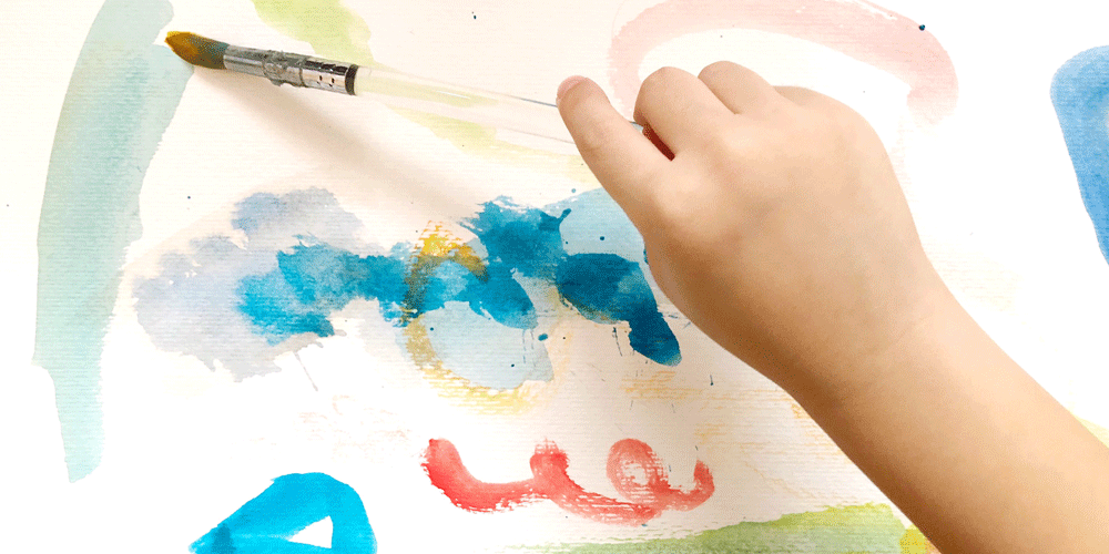 The hand of a child holding a paint brush and painting on white paper.