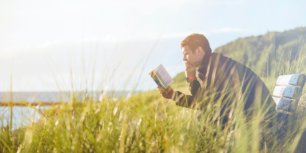 Man reading on a bench surrounded by tall grass, water and grassy hills.