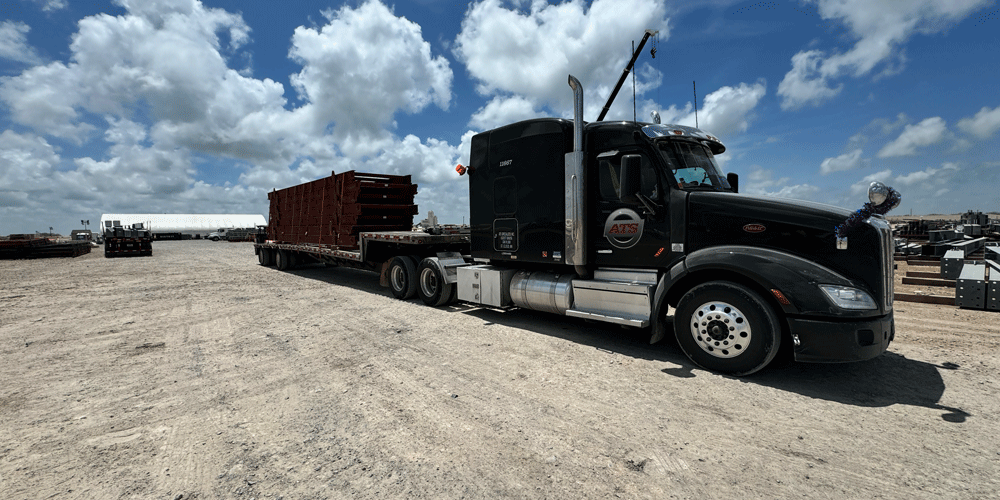 Black semi-truck with a step-deck trailer parked  in a dirt lot. The sky is cloudy. 