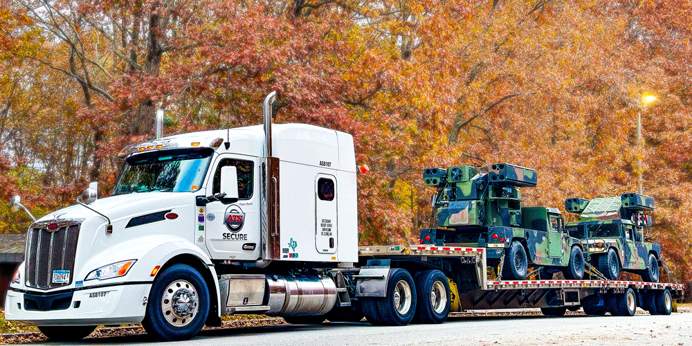 An ATS Secure truck hauling military equipment. The truck is parked in front of beautifully colored autumn leaves.