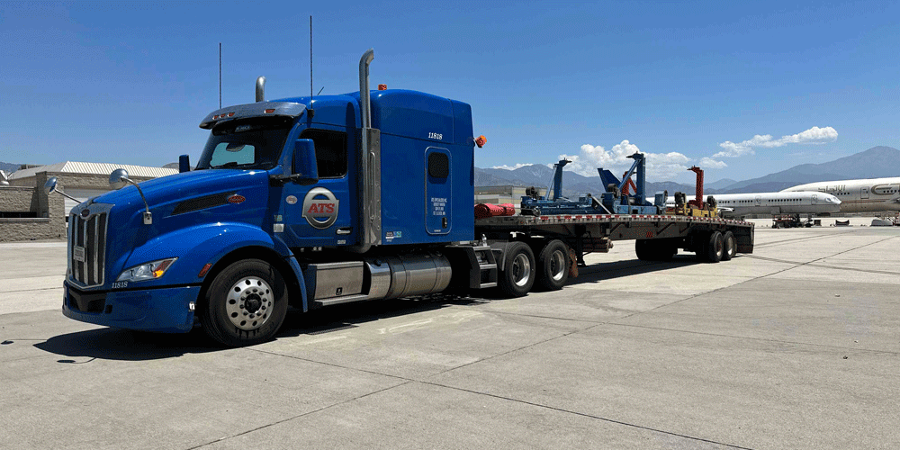 ATS truck with flatbed trailer parked at an airport with mountains in the background.