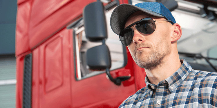 Truck driver in sunglasses and a blue trucker hat standing in front of his truck.