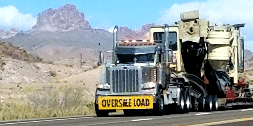 Semi-truck hauling load of machinery. An oversize load banner in yellow is on the front grill.