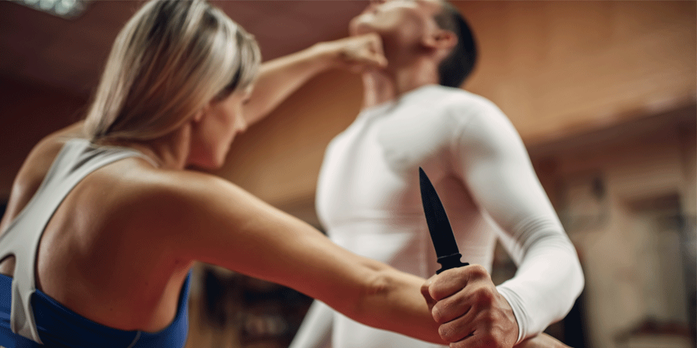 Man in woman in self defense class. The man holds a knife as the woman moves to block the blade and punch him in the throat.