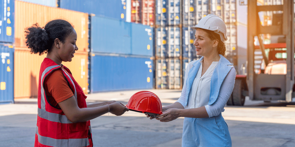 Woman in blue suit handing a woman in a safety vest a red safety helmet.
