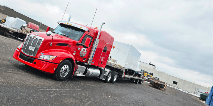 Red ATS flatbed truck with a step-deck trailer hauling a large white square-shaped piece of equipment. The truck is parked at a shipper site.
