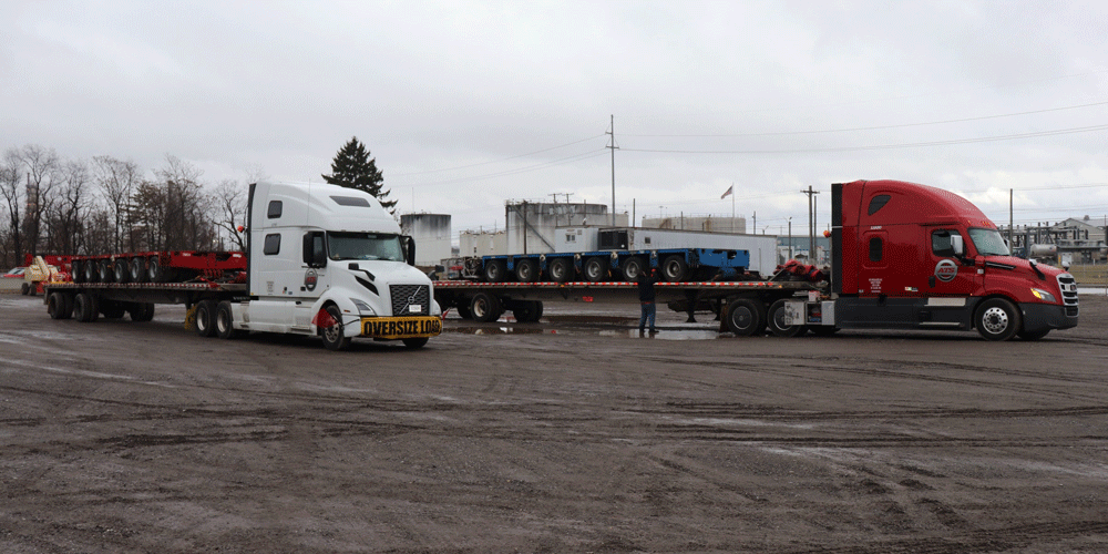 Two semi-trucks with flatbed loads parked in a dirt lot.